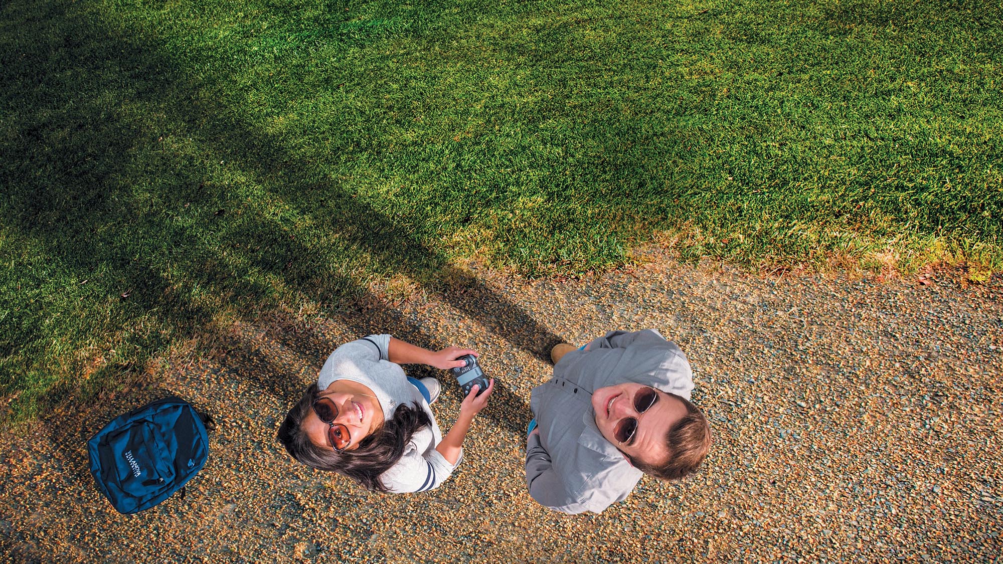 Looking up wearing sunglasses, grad students Julia McMahon and Nick Zoccoli stand in a gravel lot in Madagascar flying drones