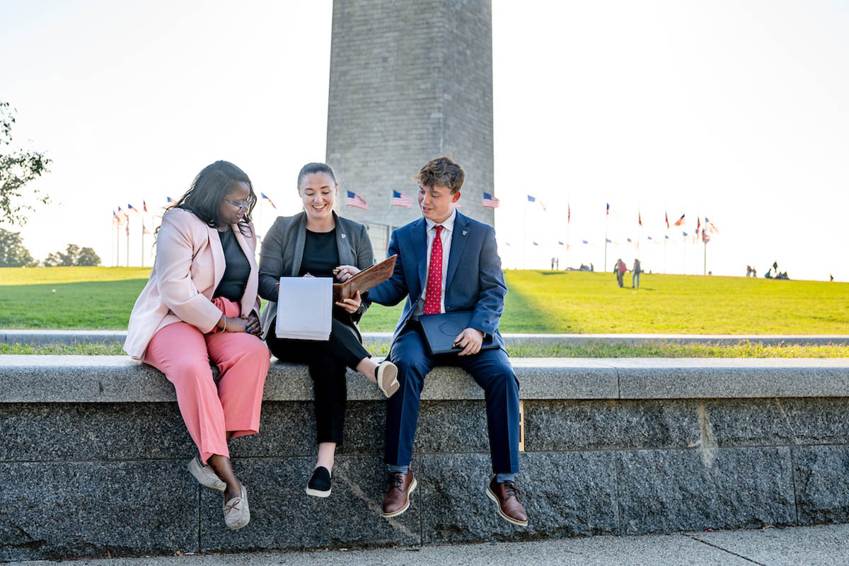 Three students sitting on a wall in Washington D.C. with the National Monument in the background.