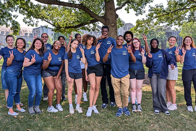group of students standing outside in matching T-shirts holding up the "V" sign with their fingers