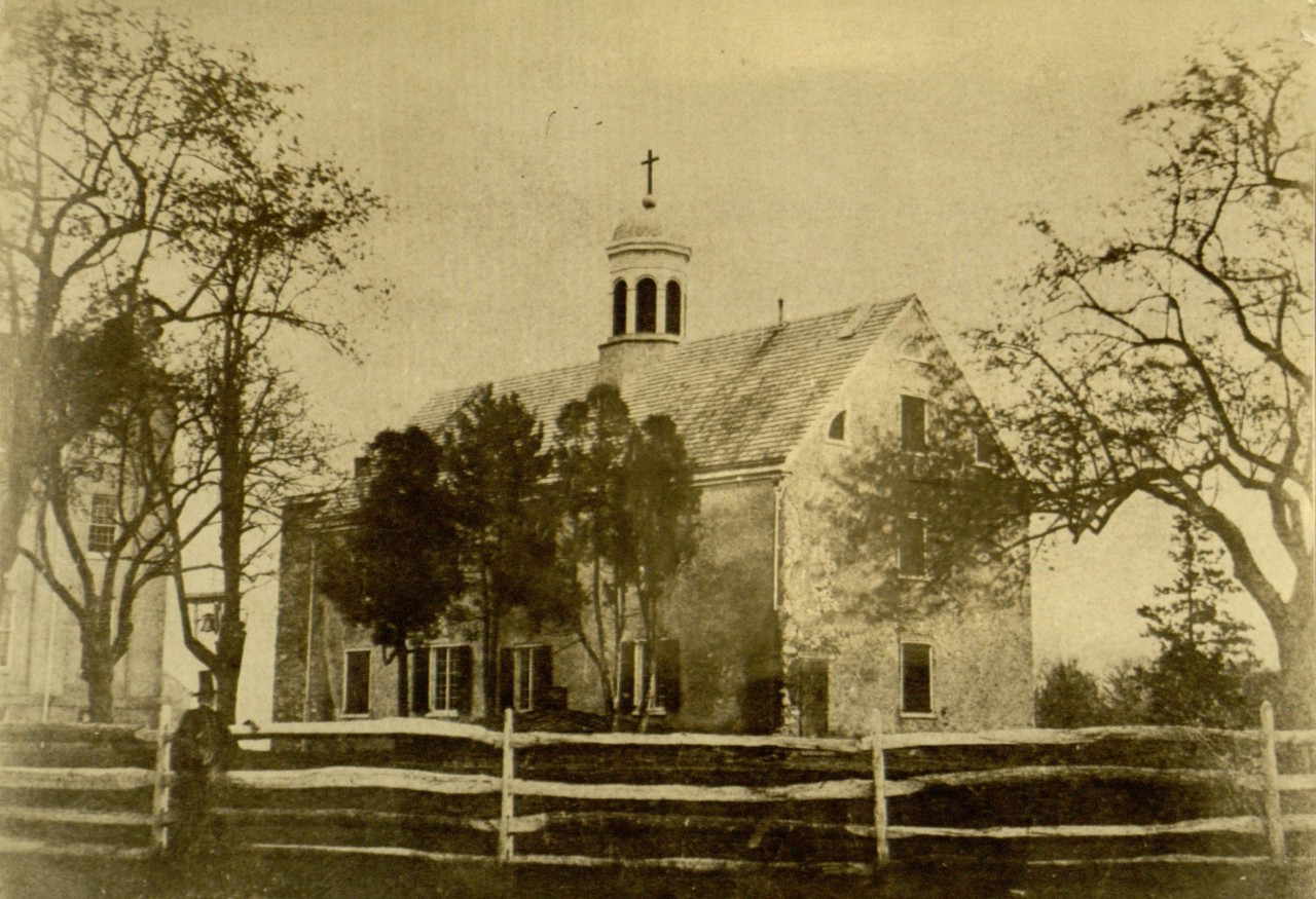 In this 1856 photo of the original chapel on campus, the Sister Bell can be seen affixed to the tree to the left of the building, just above the top hat of the gentleman near the fence.