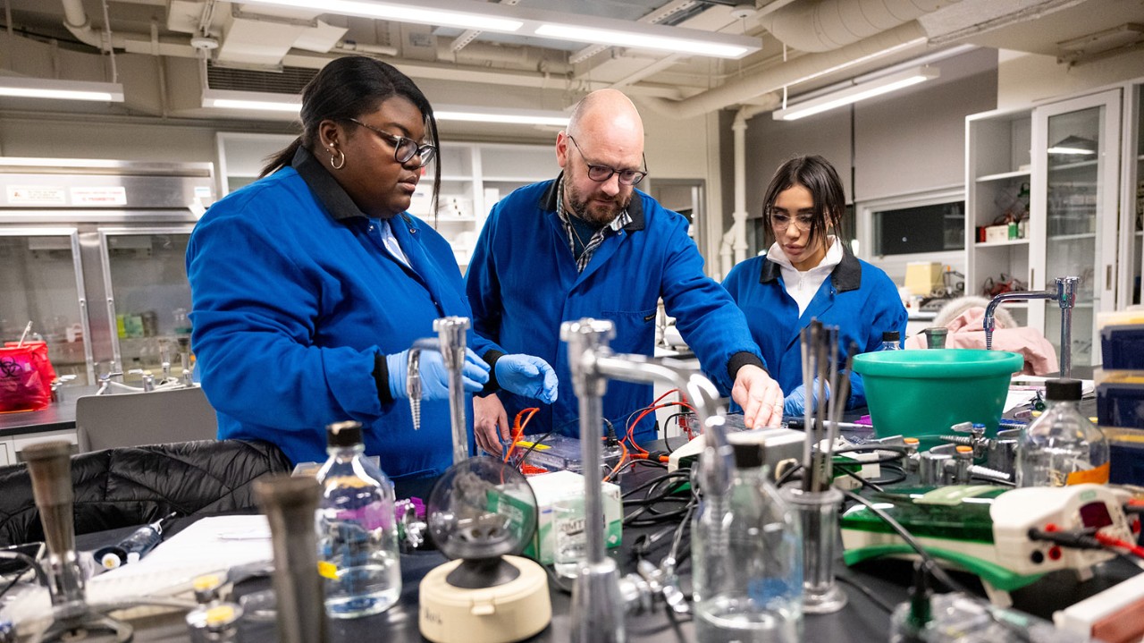 professor in biology lab with graduate students, all wearing blue lab coats and protective glasses, beakers and instruments in foreground
