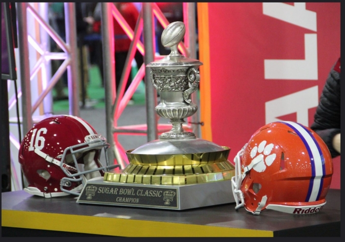 2018 Sugar Bowl Trophy Silver football trophy flanked on either side by football helmets. One red, one orange.