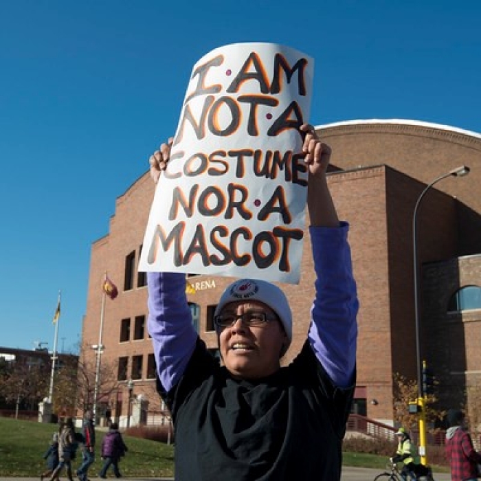 Native American woman holding sign reading I am not a costume nor a mascot standing in protest in front of the Washington football team's stadium