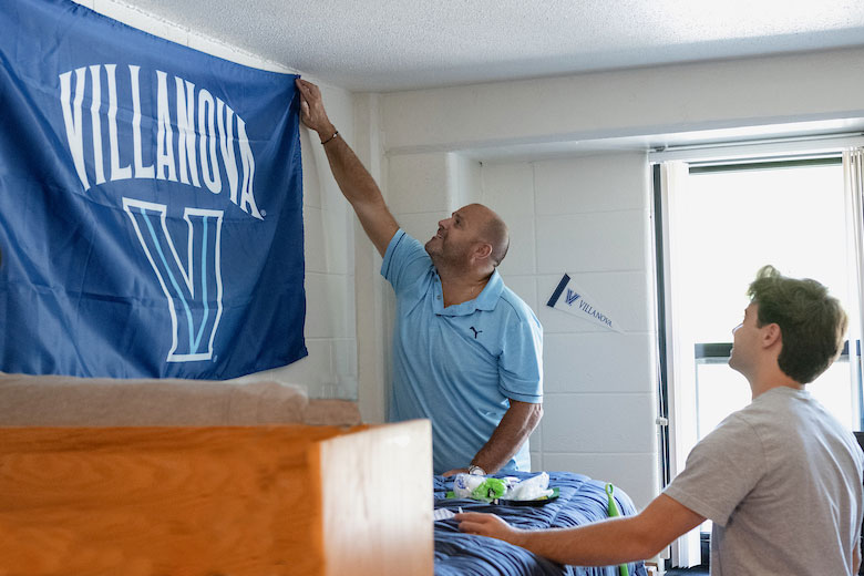 Father hanging a Villanova flag in dorm room with son watching.