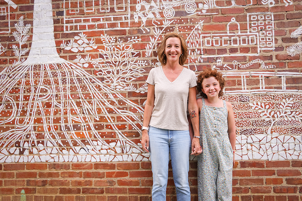 Woman and young girl smiling in front of a mural of tree roots and a building