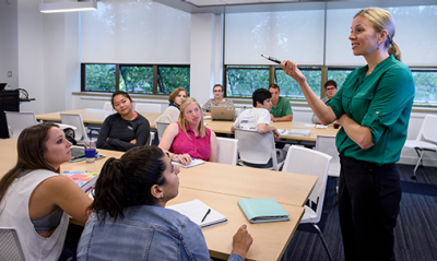Standing female faculty member speaking to students who are sitting at a conference table