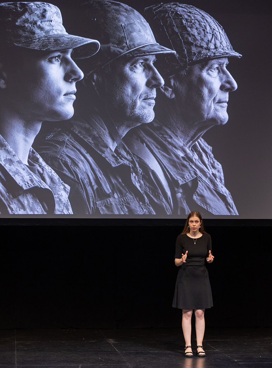 Nastia Chulkova on stage in front of her 3MT slide, which is a photo of three soldiers of different ages