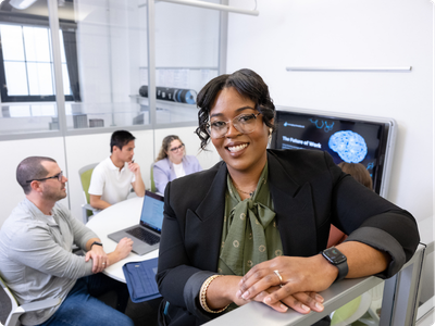 woman looking at camera in the foreground with a group of employees viewing a presentation in the background