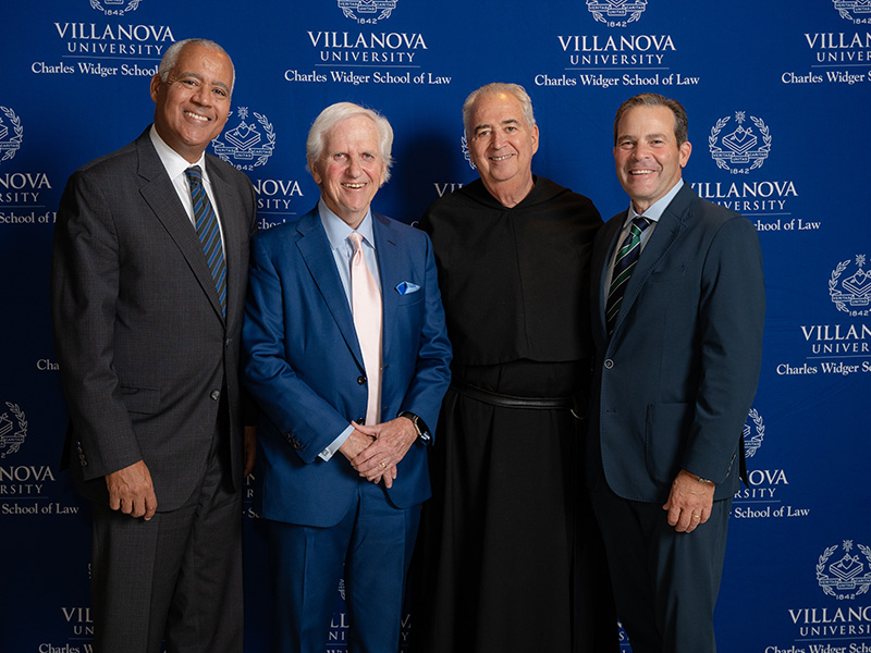 Four people pose for a group photo in front of a blue Villanova University step-and-repeat backdrop.