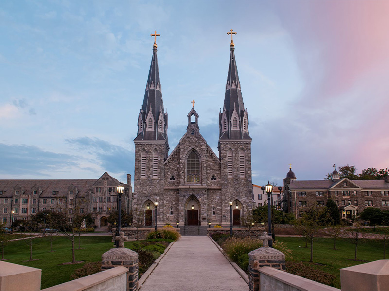 St. Thomas of Villanova Church, viewed from a central campus walkway at dusk, with surrounding lawns and campus buildings.
