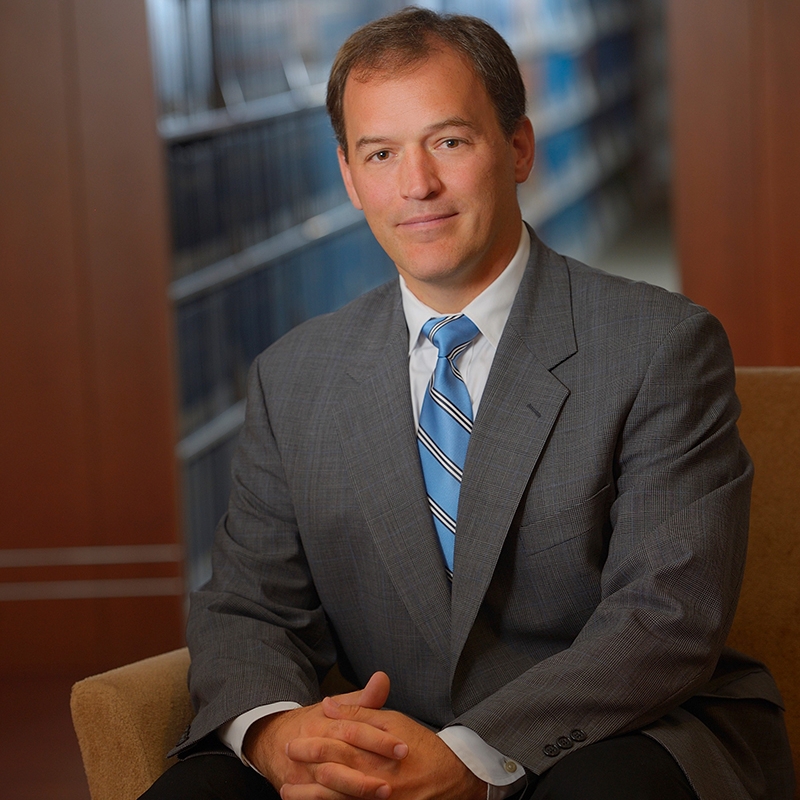 Professional portrait of a person seated in a chair, wearing a gray suit and blue striped tie, with bookshelves blurred in the background
