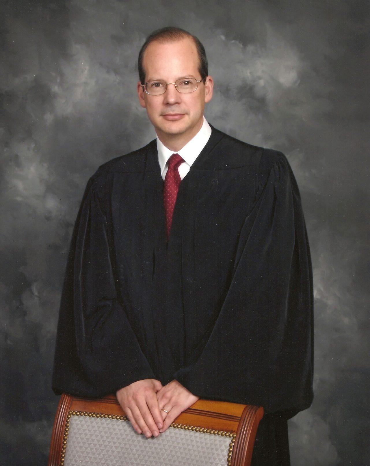 Headshot of a man in judicial robes stands behind a chair and smiles