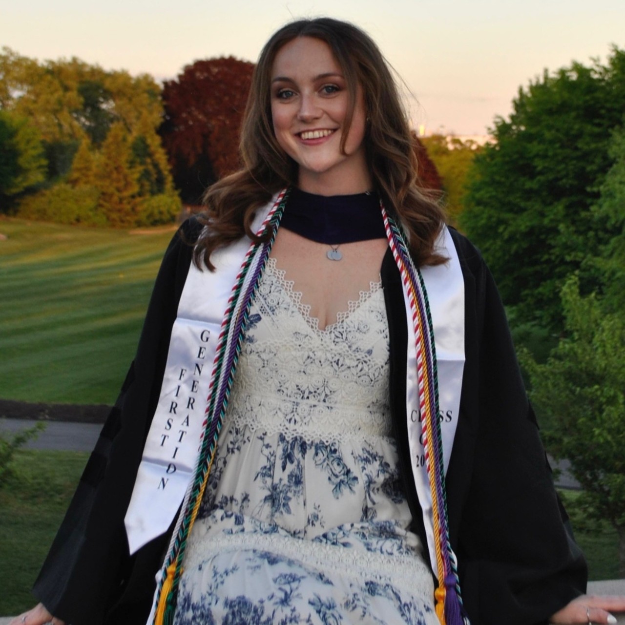 Photo of a woman in a white dress and a graduation gown standing in front of trees and grass