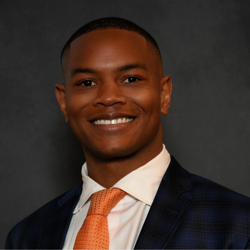  Headshot of a man in a suit with an orange tie against a gray background
