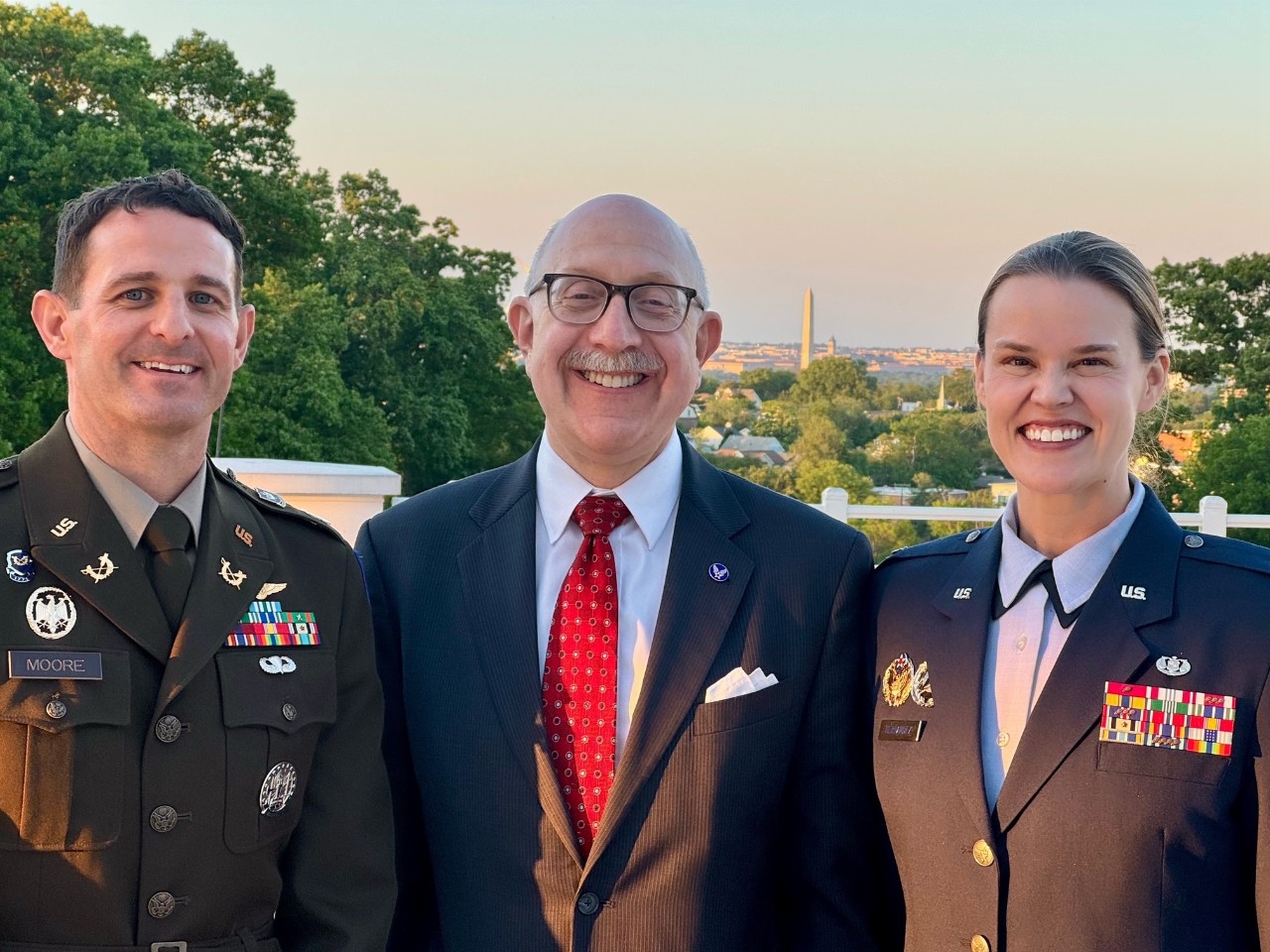 Two people in military uniforms and a man in a suit stand smiling in front of trees and buildings.
