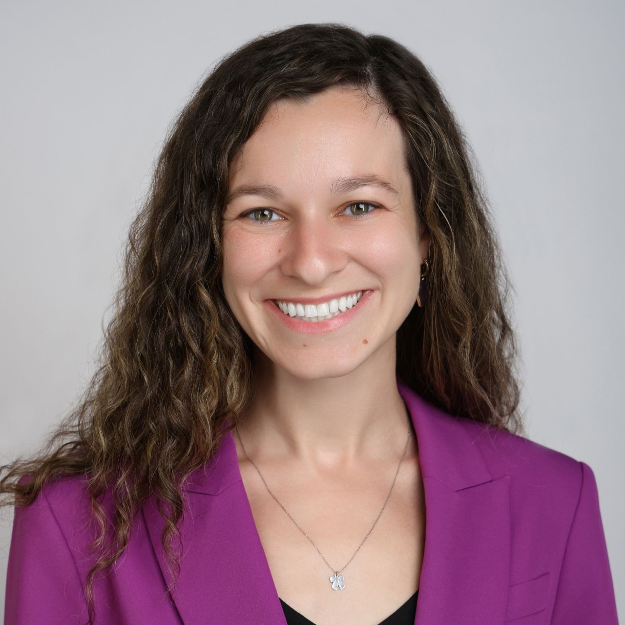 A woman in a purple blazer smiles in front of a gray background.