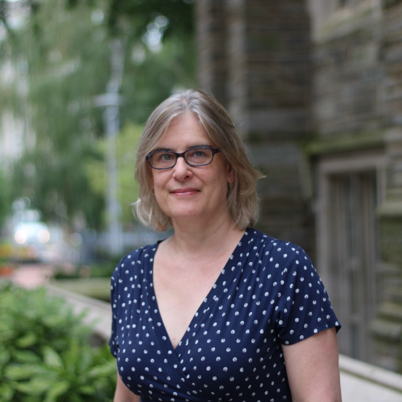 A woman in a navy polkadot shirt stands in front of a gray stone building.