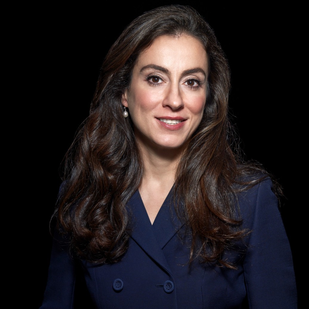 A woman in a navy blazer smiles in front of a black background.