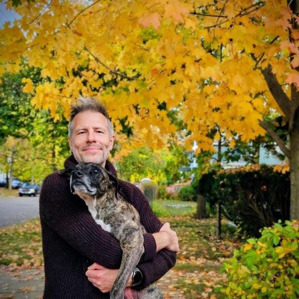 A man holds a brown dog and smiles in front of a tree with yellow and orange leaves.