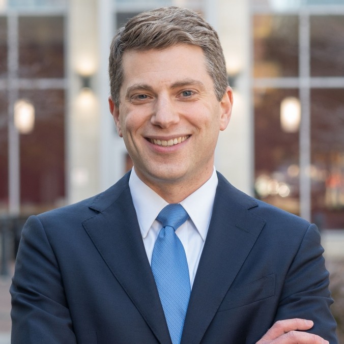 A man in a navy suit smiles in front of a white wall with windows.