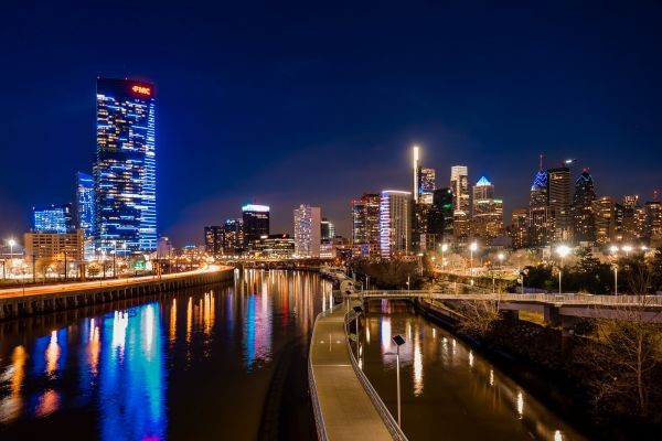 Nighttime view of the Philadelphia skyline reflected in a river, with illuminated skyscrapers, a waterfront walkway, with colorful lights mirrored on the water.