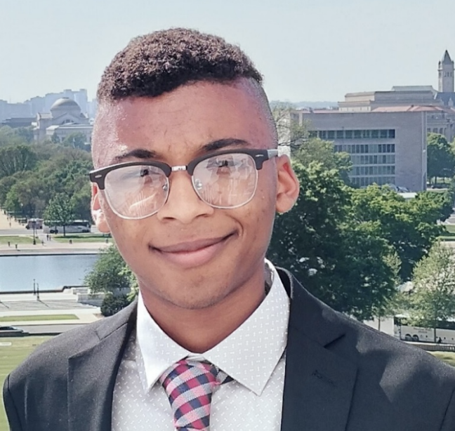 Headshot of a man wearing glasses and a tie, standing in front of a background of trees and buildings
