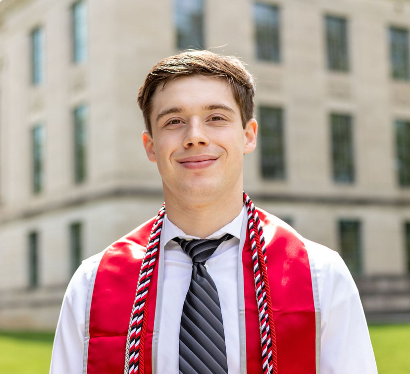 Headshot of a man wearing a white button down, tie and graduation regalia, standing in front of a blurred background of a building