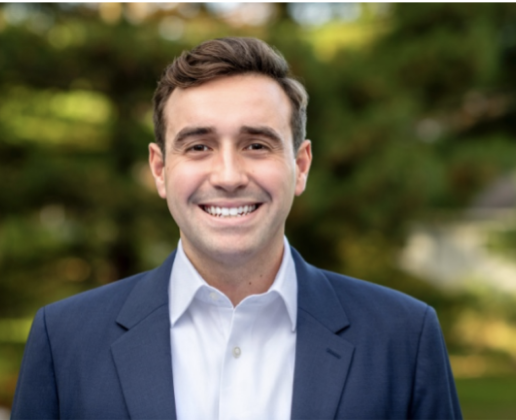 Headshot of  a man wearing a blue blazer standing in front of a blurred background of greenery