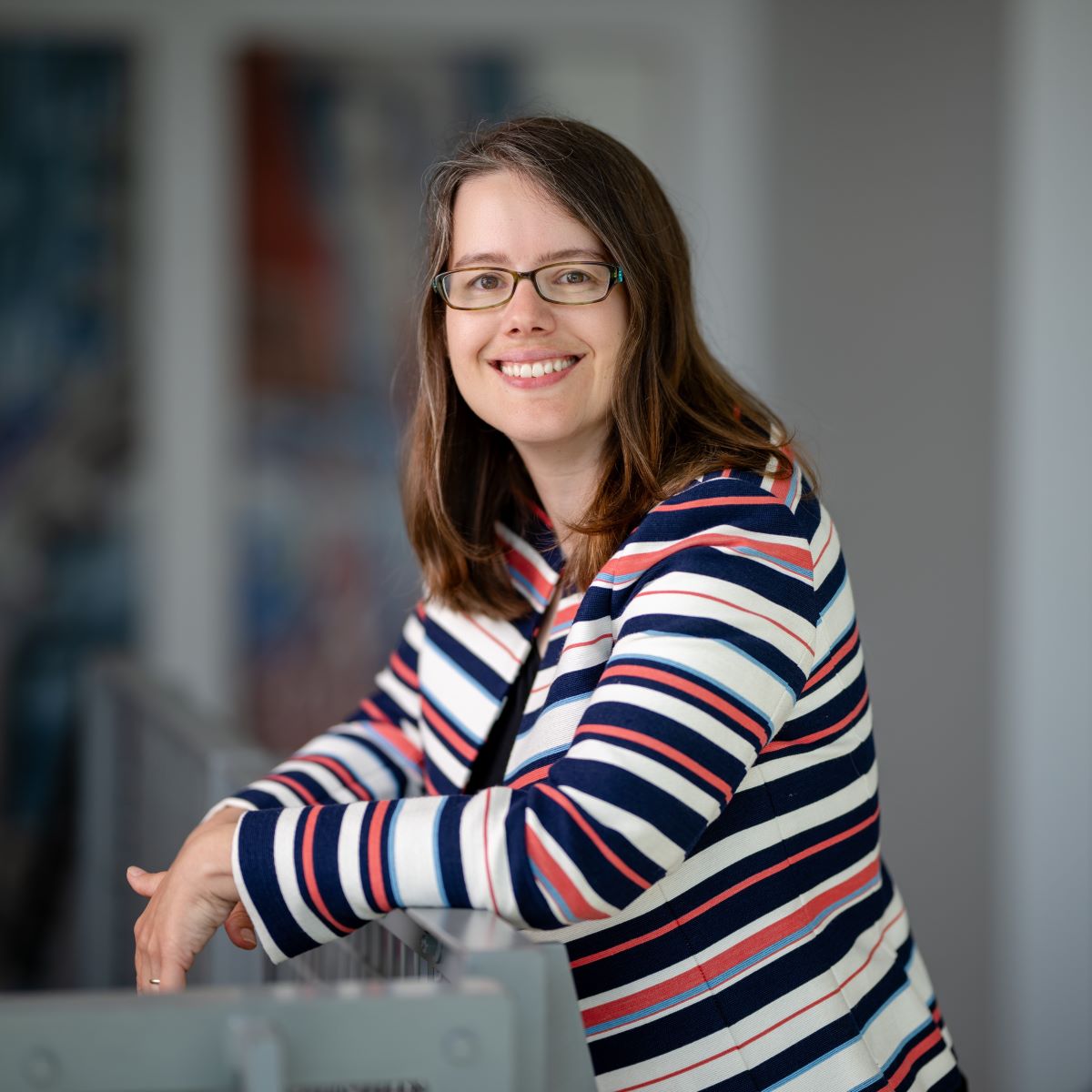 Professional headshot of a person wearing glasses and a navy, red, and white striped blazer, leaning on a railing indoors.