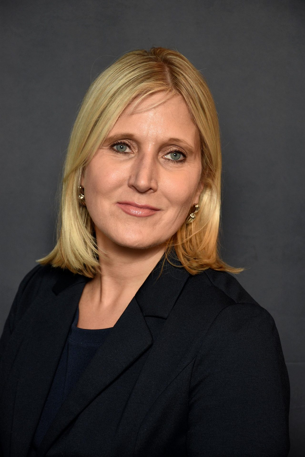 Headshot of  woman with blonde hair wearing a black blazer posed in front of a gray background