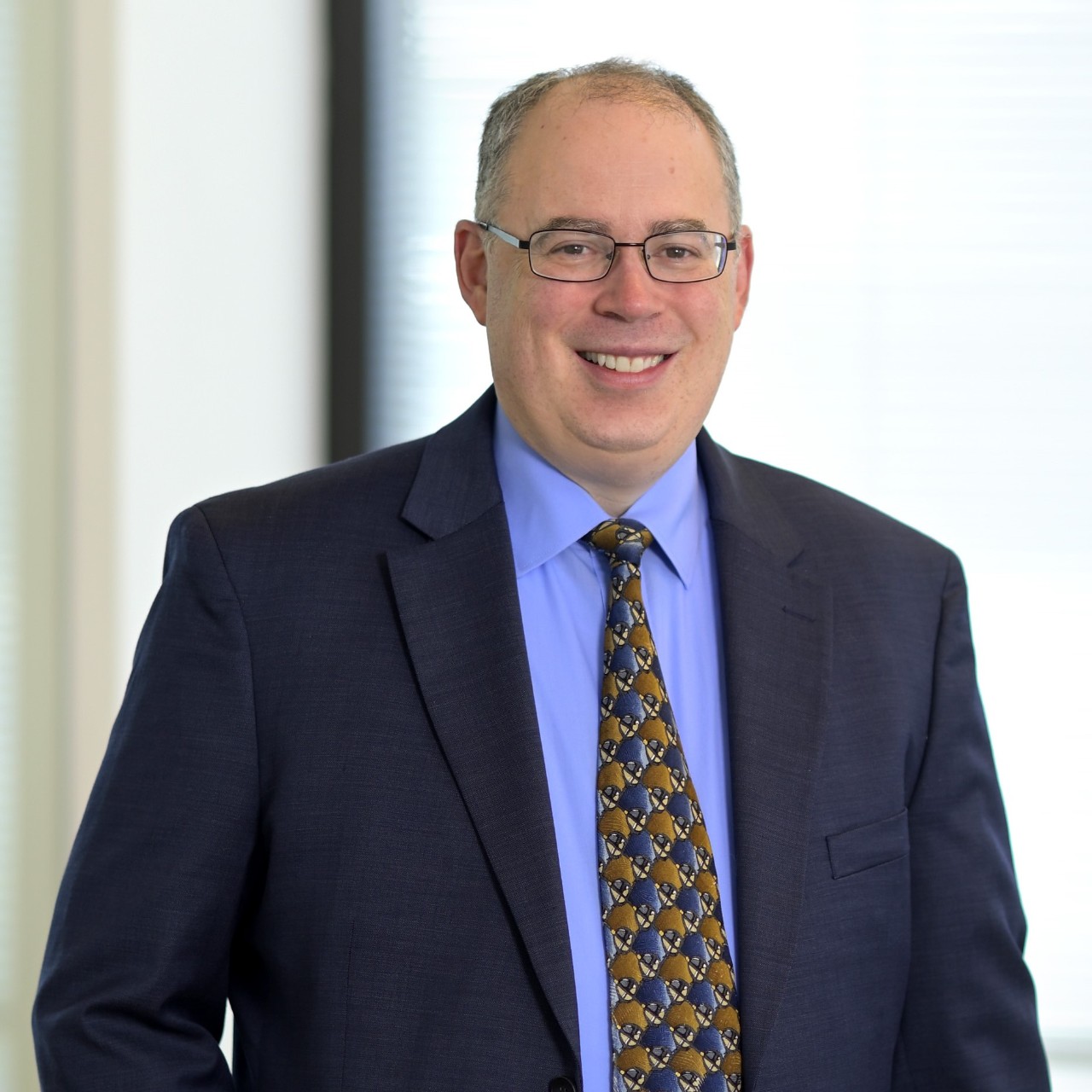 Professional portrait of a person wearing glasses, a navy suit, a blue dress shirt, and a patterned tie, standing indoors against a softly blurred background