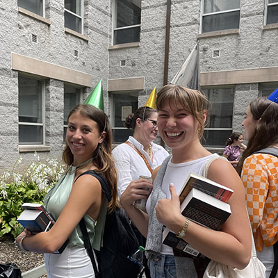 Humanities alumni holding books outside and wearing party hats.
