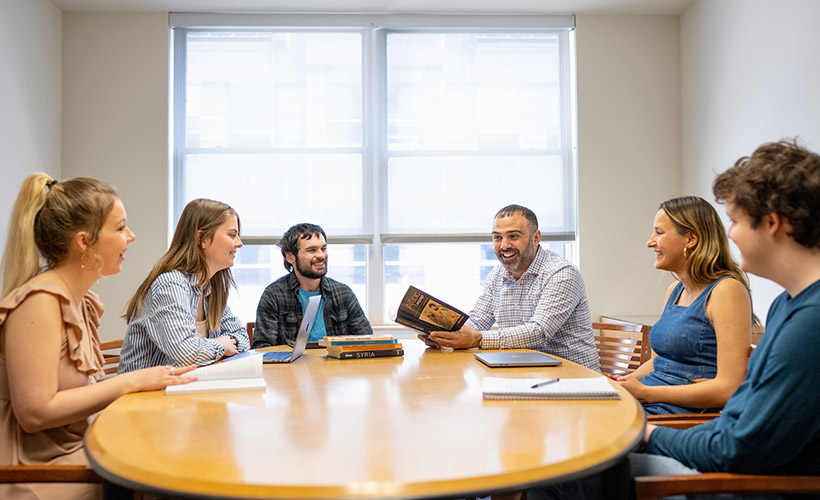 Students sit around a table with a professor in discussion.