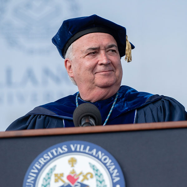 Father Peter in academic regalia stands behind a podium at Commencement
