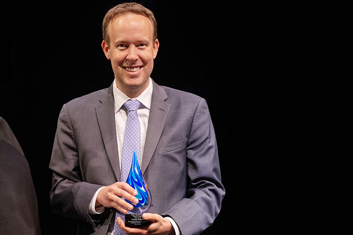 Timothy Hoffman wears a gray suit and smiles while holding the Steve Merritt Award