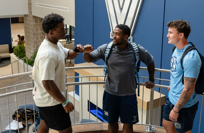 Three male Villanova students on the staircase in the newly renovated Connelly Center, with two exchanging a fist bump.