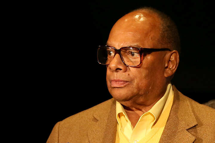 George Raveling, Villanova basketball legend and Hall of Fame coach, in a headshot wearing glasses, a yellow shirt, and a brown suit jacket against a black background, looking to the side.