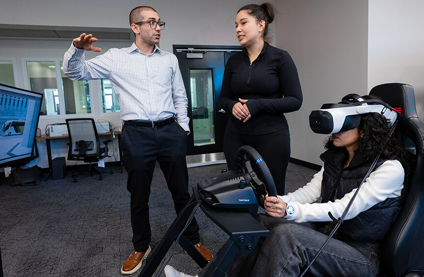 A professor works with graduate students in a lab; one student wears a VR headset and uses a steering wheel to drive a transportation simulator.