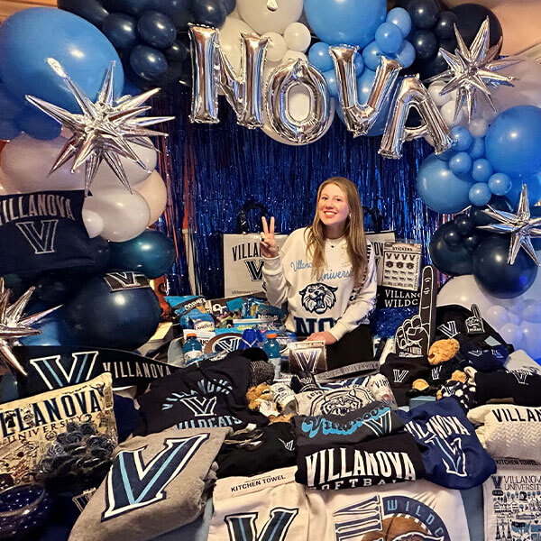 Young woman celebrating her admission to Villanova, sitting on her bed surrounded by Villanova gear including T-shirts, with silver balloons spelling ‘NOVA’ and additional balloons in Villanova’s blue and white colors.
