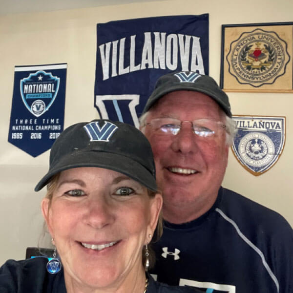Couple standing in front of a wall decorated with Villanova banners and flags, both wearing blue Villanova ‘V’ baseball caps.