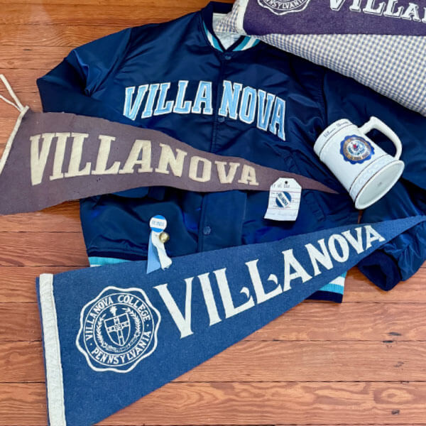 Villanova blue sweatshirt laid out on a wooden floor alongside three Villanova pennants and a Villanova ceramic beer mug.