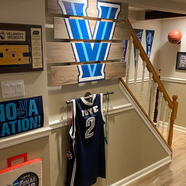 Basement room decorated with Villanova basketball memorabilia, including pennants, a wooden ‘V’ wall hanging, a basketball, a Foye No. 2 jersey, and other Villanova-themed items.