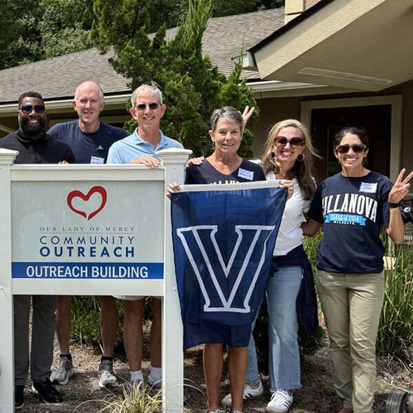 Group of six Villanova volunteers standing outdoors, holding a Villanova ‘V’ flag and posing next to a sign that reads ‘Community Outreach.’