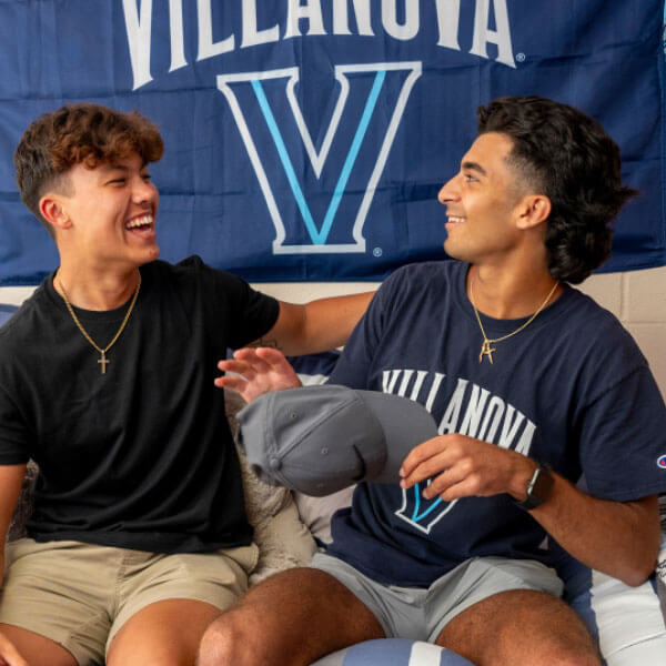 Two male students sitting together in front of a Villanova banner, smiling and enjoying each other’s company.