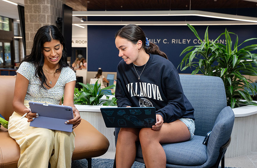 Two female Villanova students seated side by side on the ground floor of the newly renovated Connelly Center, looking at their digital screens.
