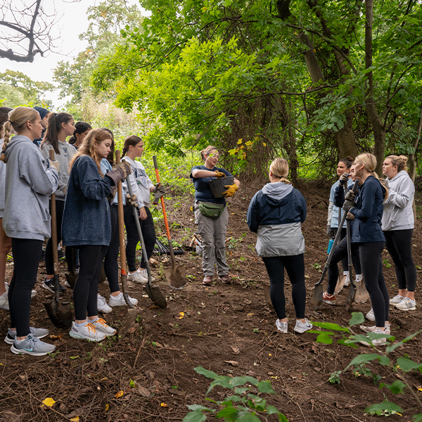 A group of Villanova volunteers with shovels stand outside on a dirt path surrounded by trees