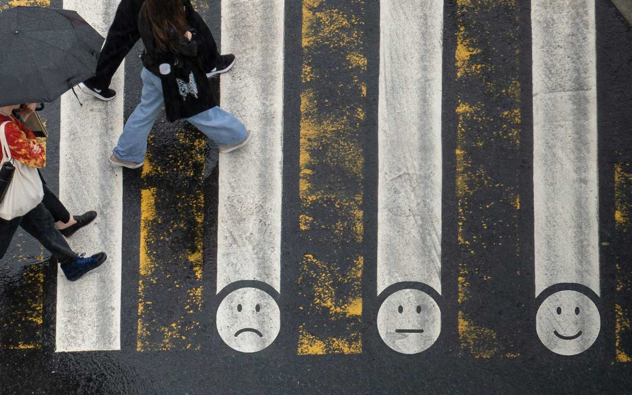 Aerial view of people crossing a crosswalk with alternating faded yellow and white lines. Three of the white lines feature faces representing sad, neutral, and happy emotions, similar to a pain scale.