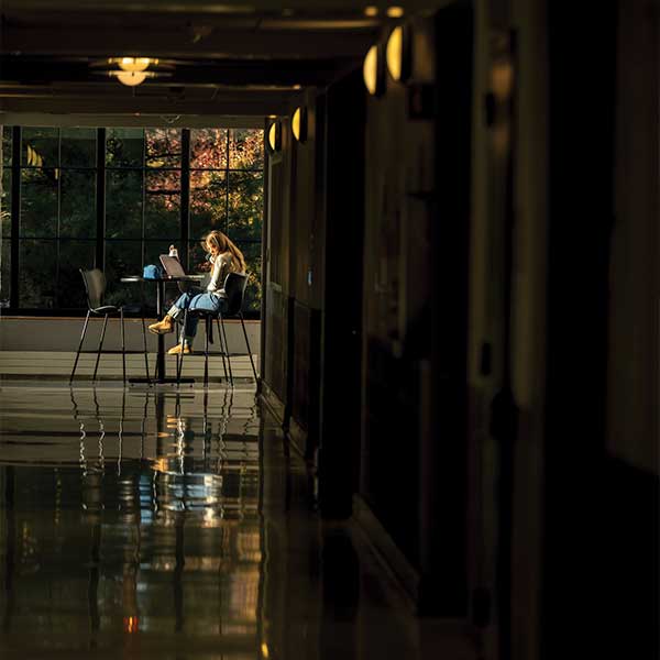 A student sits alone at a high-top table on a stool, with a large window behind her, inside a building on campus.