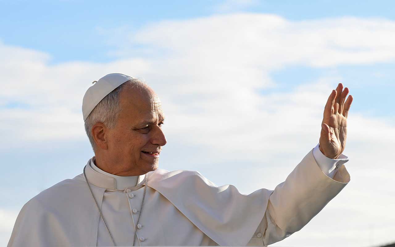 Pope Leo XIV raises a hand in greeting outdoors against a blue sky.