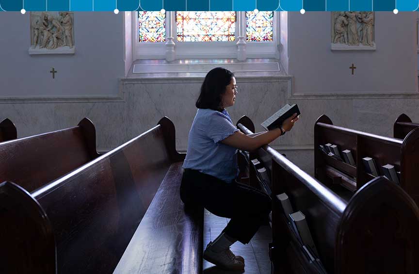 A woman sits on a chapel pew while reading from a book.
